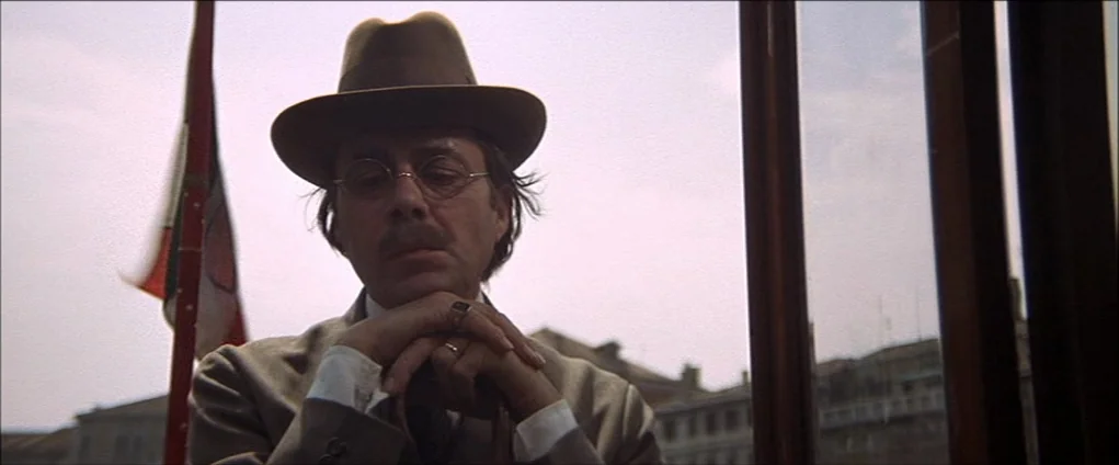 Death In Venice (1971) movie still: medium — A man stands on a terrace with an Italian flag visible in the background. He leans forward with his…