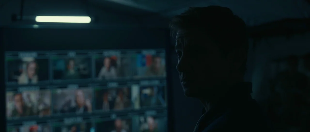 Arrival (2016) movie still: medium — A darkened room features a large wall composed of multiple live security video feeds. A man stands in the…