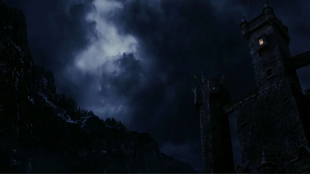 Van Helsing (2004) movie still: establishing — A dark stone castle looms against a cloudy night sky above a rocky cliffside. The structure features…