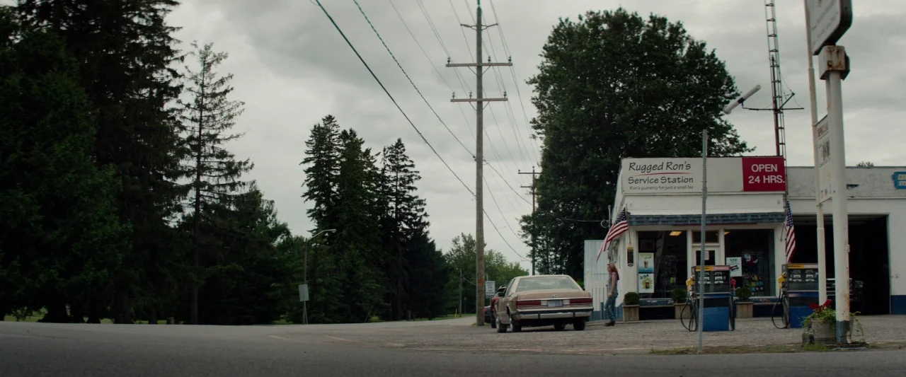 The Monster (2016) movie still: wide — A rural service station with a tan car parked by the fuel pumps. A young woman stands near the building under…