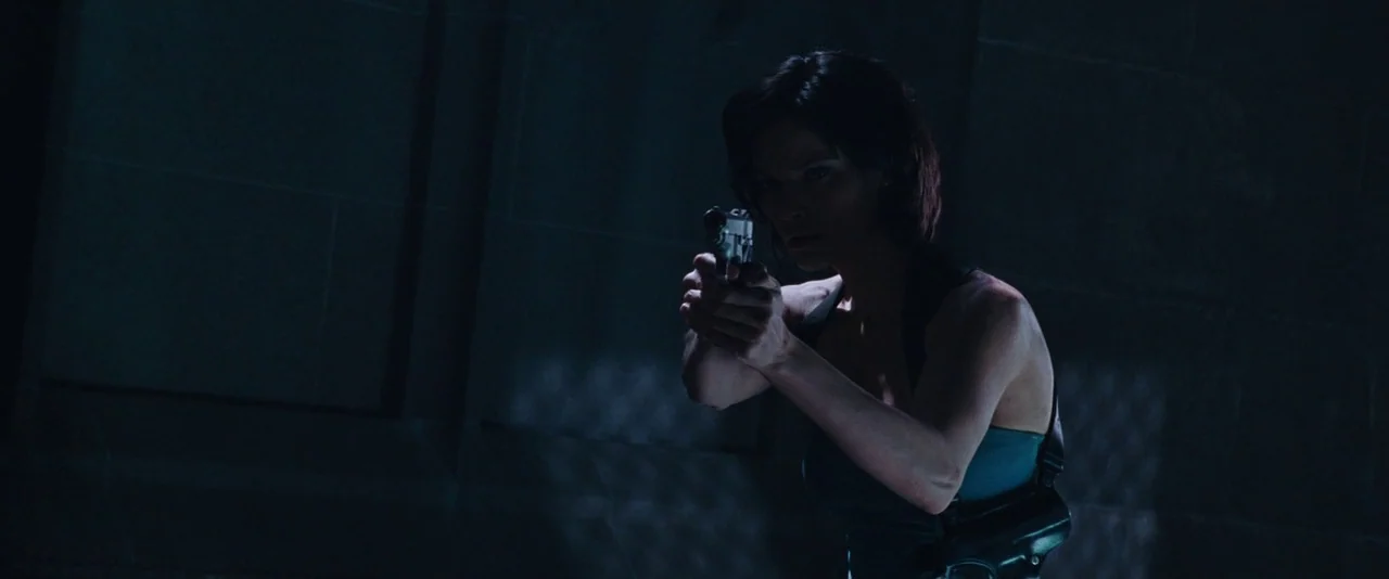 Resident Evil: Apocalypse (2004) movie still: medium — A dark, dimly lit environment features concrete walls in the background. A woman in a…