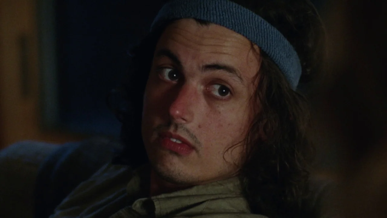 Queen of Earth (2015) movie still: closeup — A man sits in a dimly lit room with dark shadows in the background. He is wearing a blue headband and…