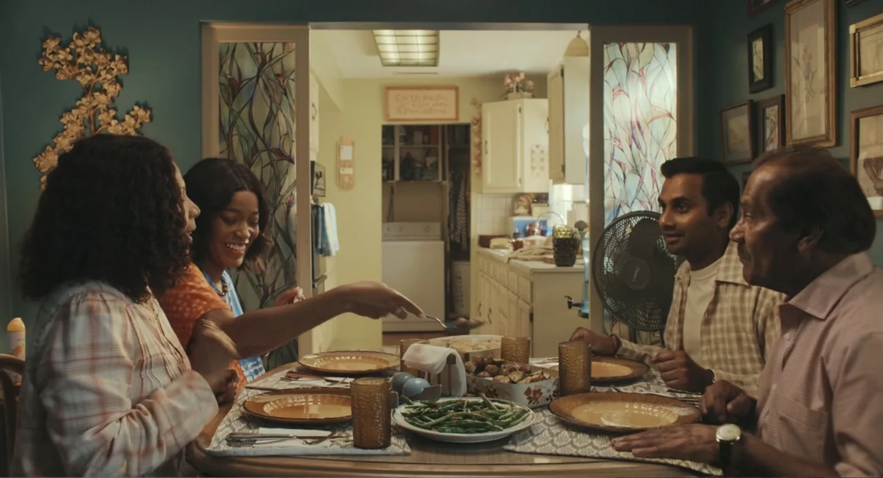 Good Fortune (2025) movie still: medium — A residential dining area has a wooden table set for four with food. Four adults sit around the table…