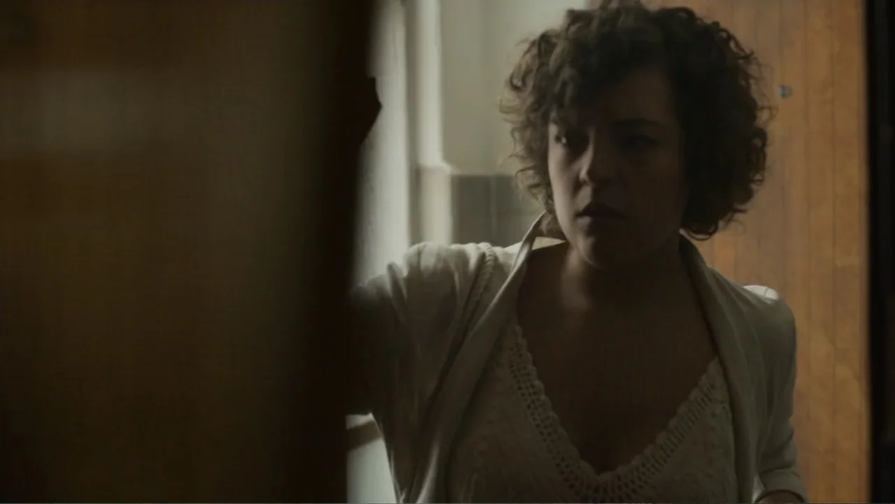 Lecciones para canallas (2022) movie still: medium — An apartment hallway with a plain wall and a partially opened wooden door frame. A young woman…