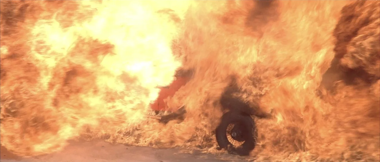 Sahara (1995) movie still: medium — A vehicle is completely consumed by a large, bright explosion on a paved surface. Thick, turbulent flames and…