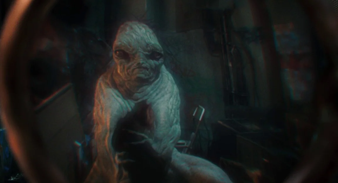Hellboy II: The Golden Army (2008) movie still: medium — A dimly lit, cluttered basement environment viewed through a small circular frame. A pale,…