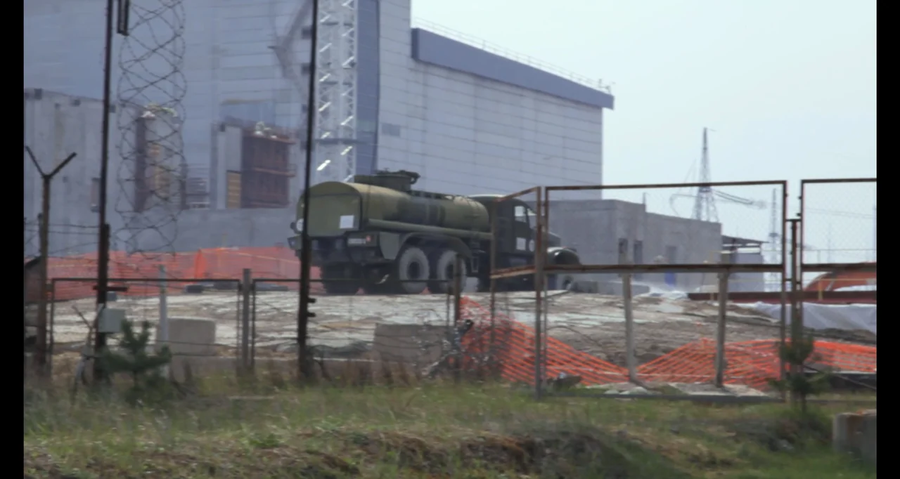 Chernobyl's Café (2016) movie still: wide — A fenced-in industrial lot surrounds a large grey structure with utility power lines nearby. A…