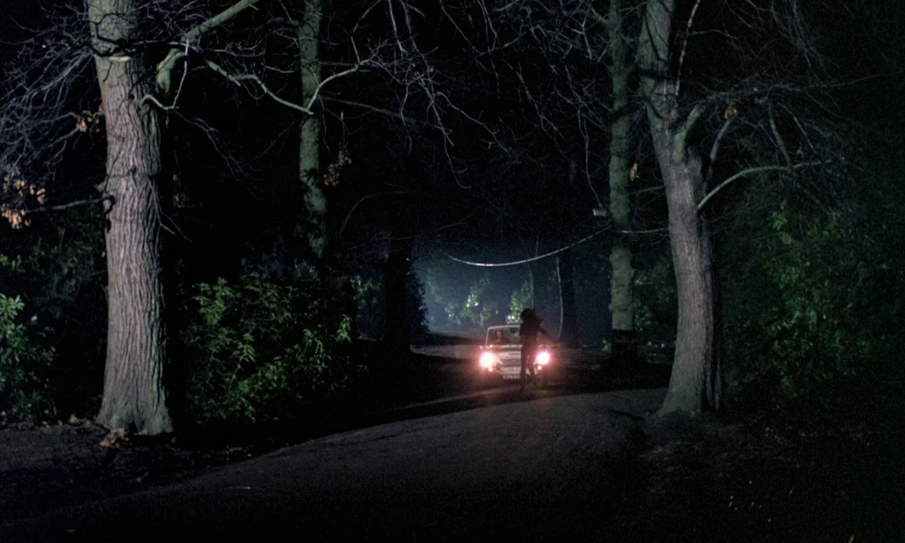 Curse of the Crimson Altar (1968) movie still: wide — A dark road winds through dense trees under a moonless night sky. A white car remains parked…