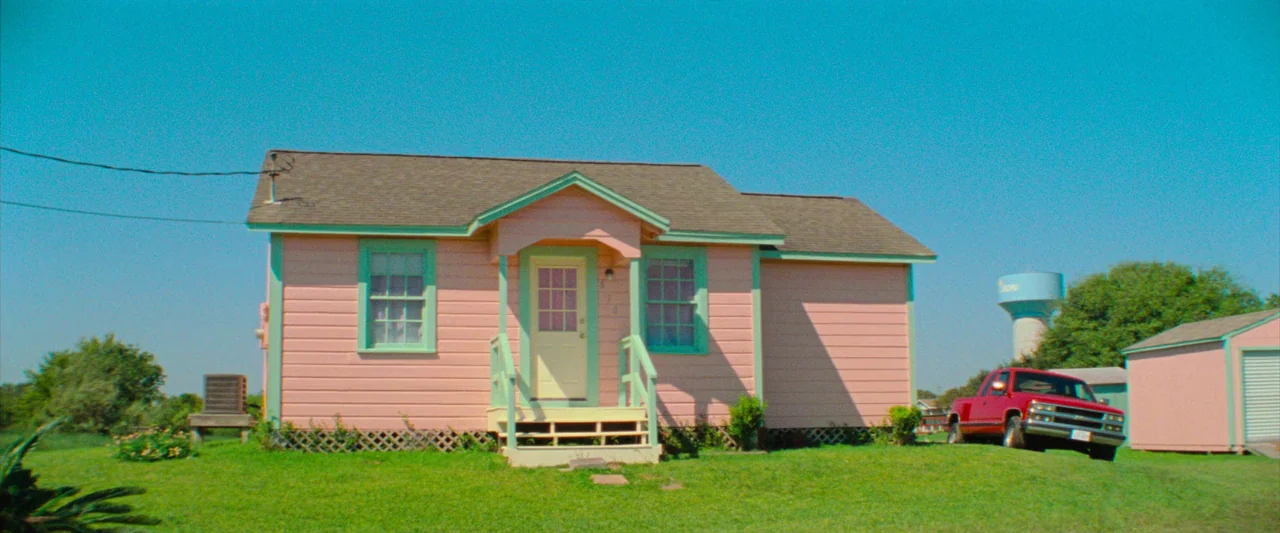 Red Rocket (2021) movie still: establishing — A single-story house painted light pink with mint green trim sits on grass. A bright red pickup truck…
