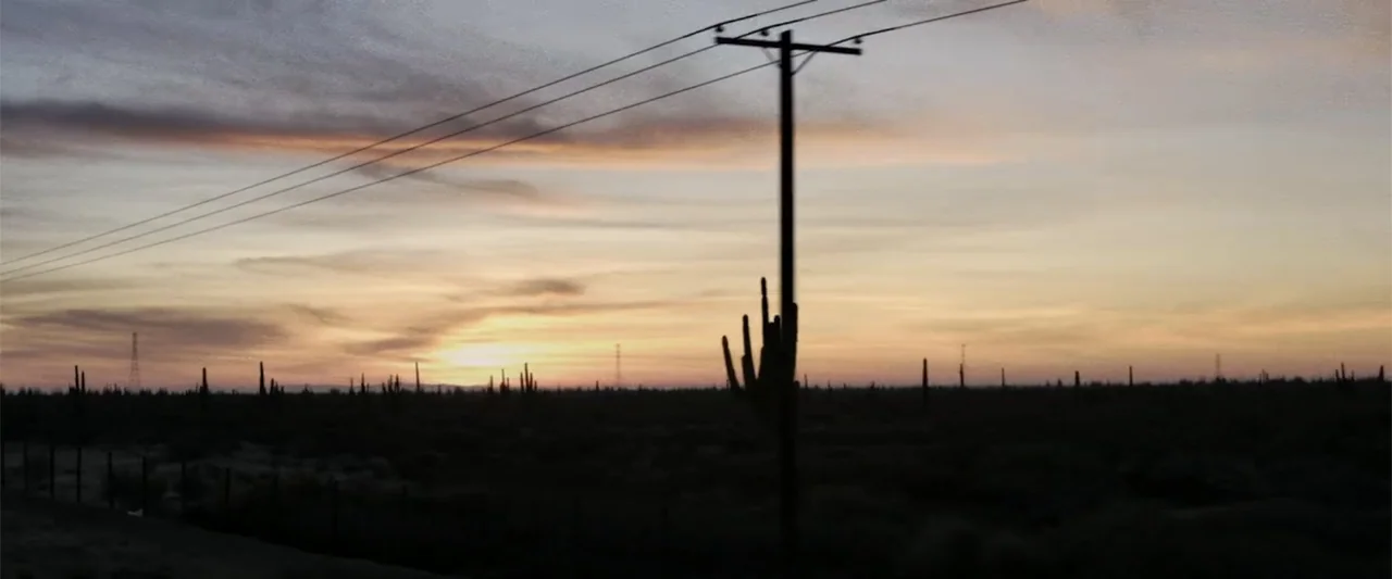 At Dawn (2013) movie still: establishing — A vast desert landscape features silhouetted cacti and a prominent utility pole. The sun sets low on the…
