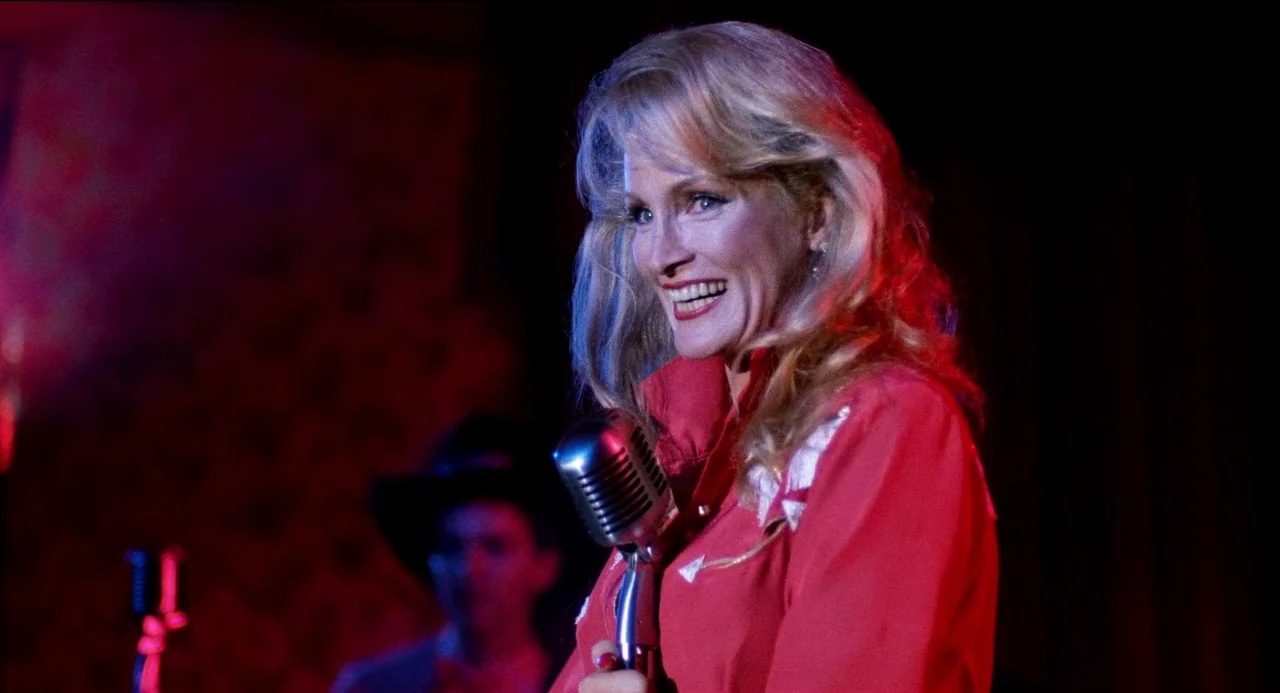 Desert Hearts (1985) movie still: medium — A dimly lit stage features a blonde woman in a red western shirt. She sings into a chrome microphone…