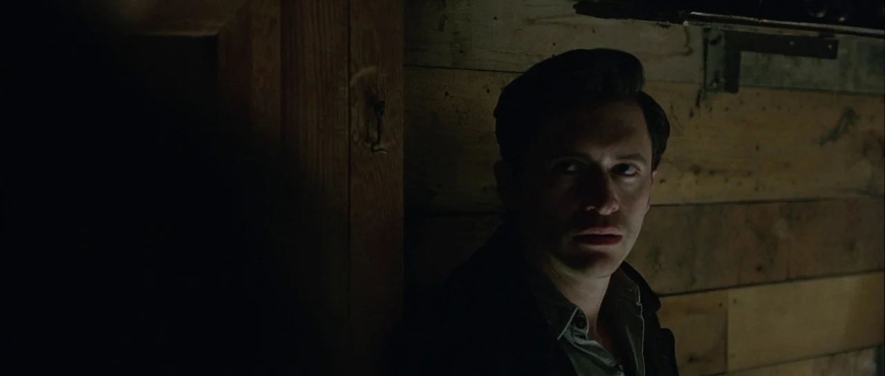 Capote (2005) movie still: closeup — A dimly lit interior with rough wooden plank walls and a dark corner. A man in a collared shirt looks toward…