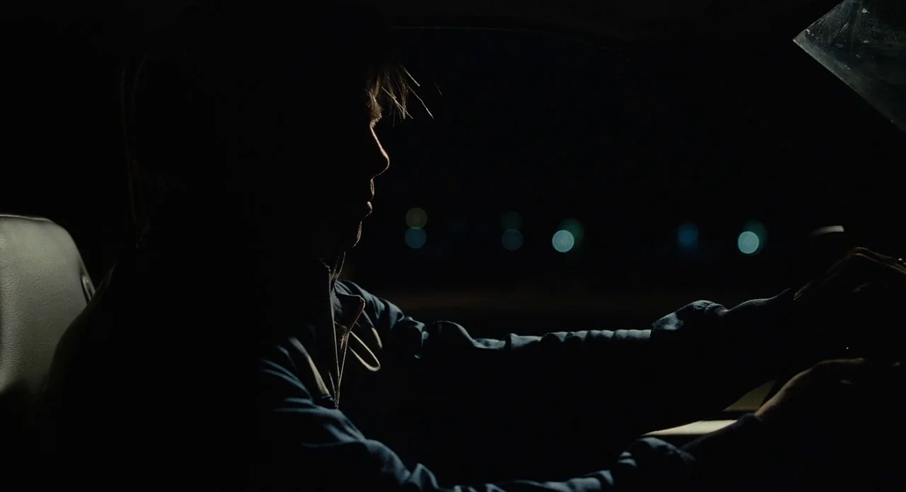 Moneyball (2011) movie still: medium — A man sits in the driver's seat of a dark car at night. He is shown in silhouette with bokeh street lights…