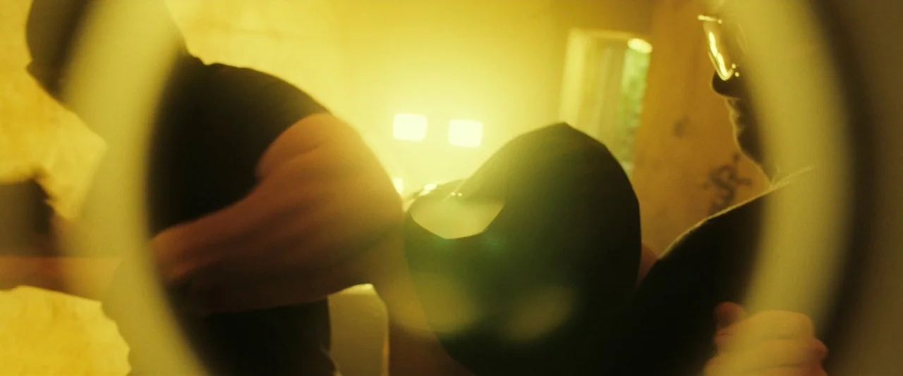 The Report (2019) movie still: medium — A dark, dimly lit room with rough walls and a bright light source. Two men stand in the foreground obscured…