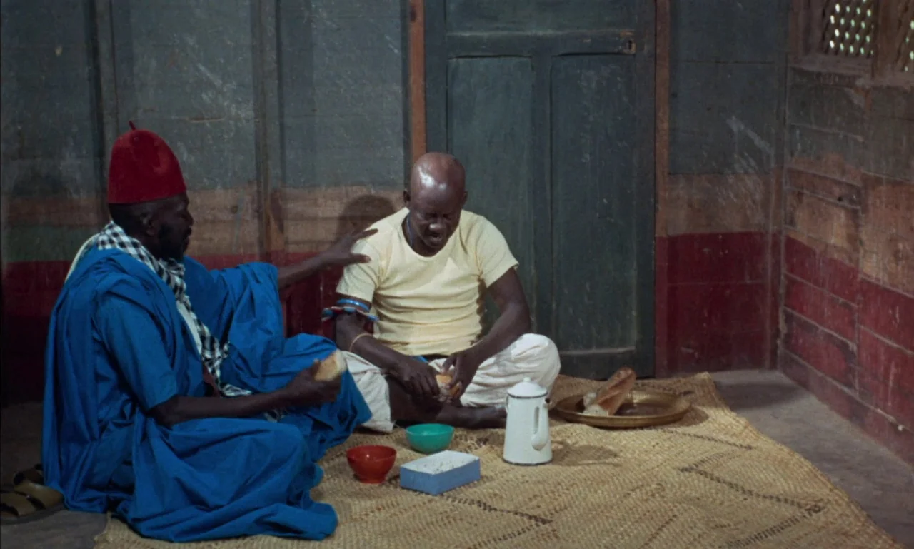 Mandabi (1968) movie still: medium — Two men are seated on a straw mat inside a room with wood paneling. They are eating bread from a tray alongside…