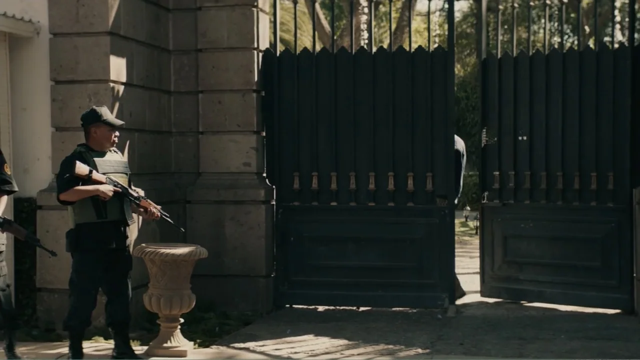 Bel Canto (2018) movie still: fullbody — An imposing black metal gate is flanked by large stone pillars in daylight. Two armed guards wearing…