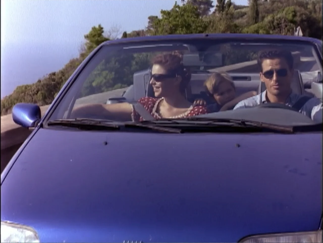 Code Name: Wolverine (1996) movie still: medium — A dark blue convertible car travels along a sunlit coastal road lined with vegetation. A man, a…