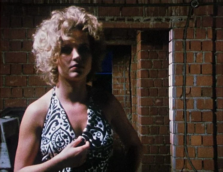 The Niklashausen Journey (1970) movie still: medium — A woman stands before a wall of exposed, dark red clay bricks. She is adjusting the strap of…