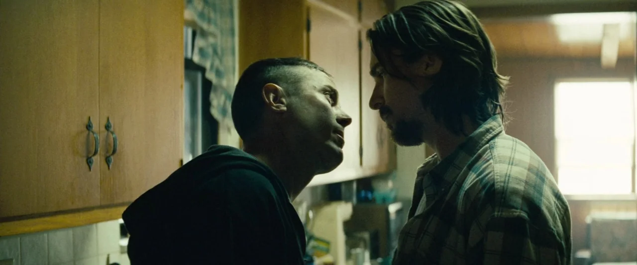 Out of the Furnace (2013) movie still: medium — A residential room features wooden cabinetry and a window partially covered by a curtain. Two men…