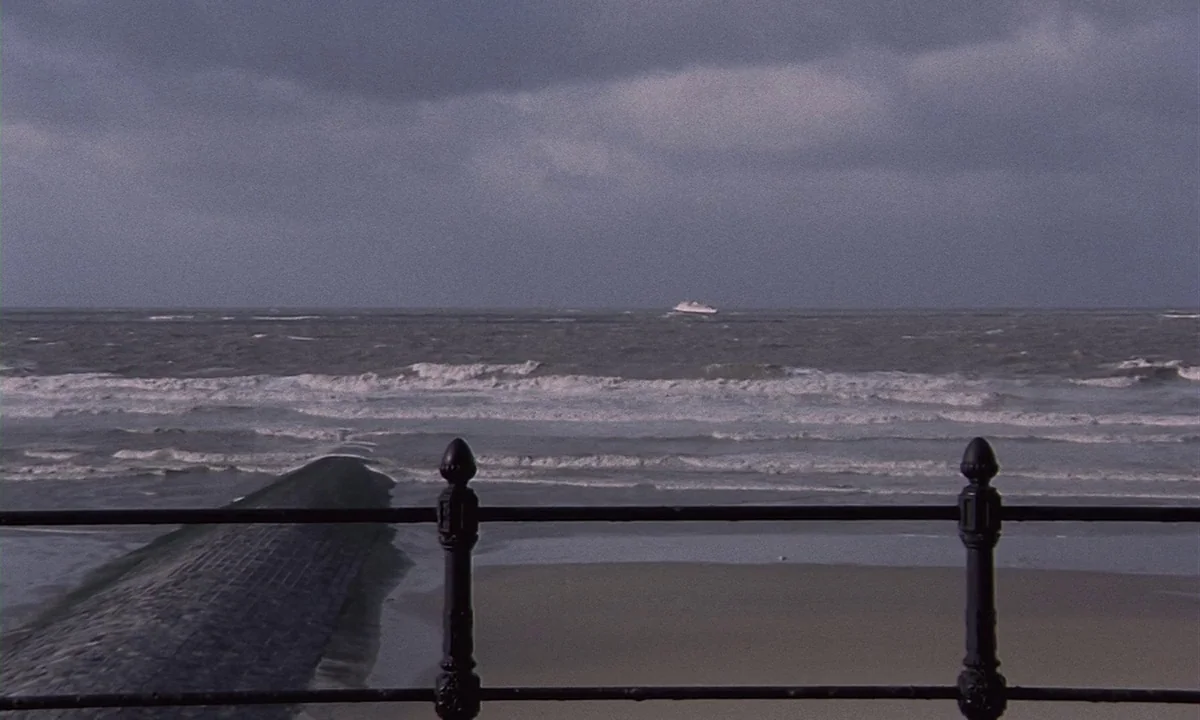 Daughters of Darkness (1971) movie still: establishing — A dark, overcast day at a beach with a stone pier stretching into surf. A small white boat…