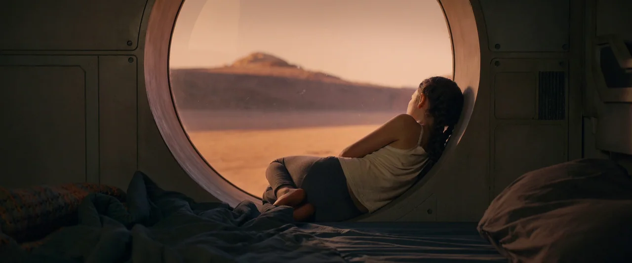 Settlers (2021) movie still: medium — A spartan spaceship interior features a large circular window looking out onto a desert. A young girl lies…