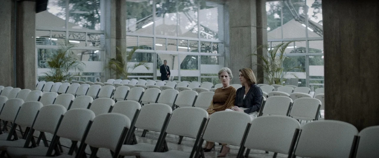 La Llorona (2019) movie still: wide — A spacious auditorium features rows of gray chairs and large floor-to-ceiling glass windows. Two women sit…