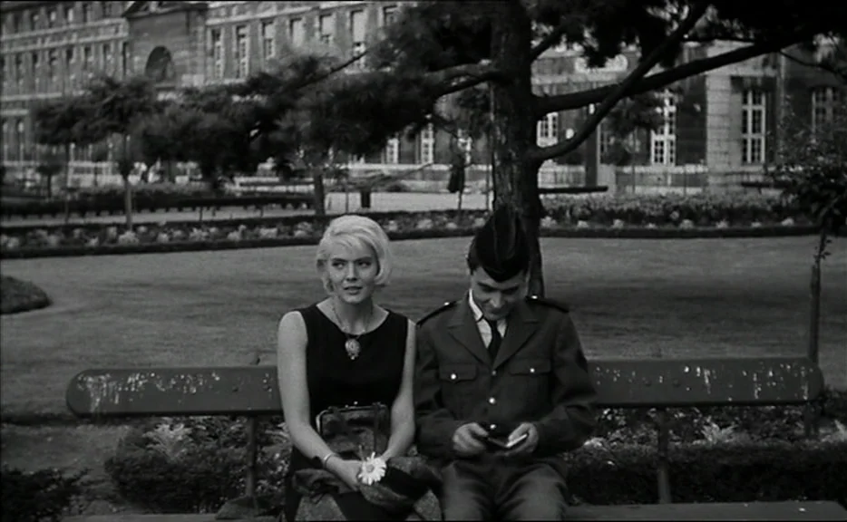 Cléo from 5 to 7 movie still: medium — A woman and a uniformed man sit together on a park bench. An old stone building and a large pine tree are…