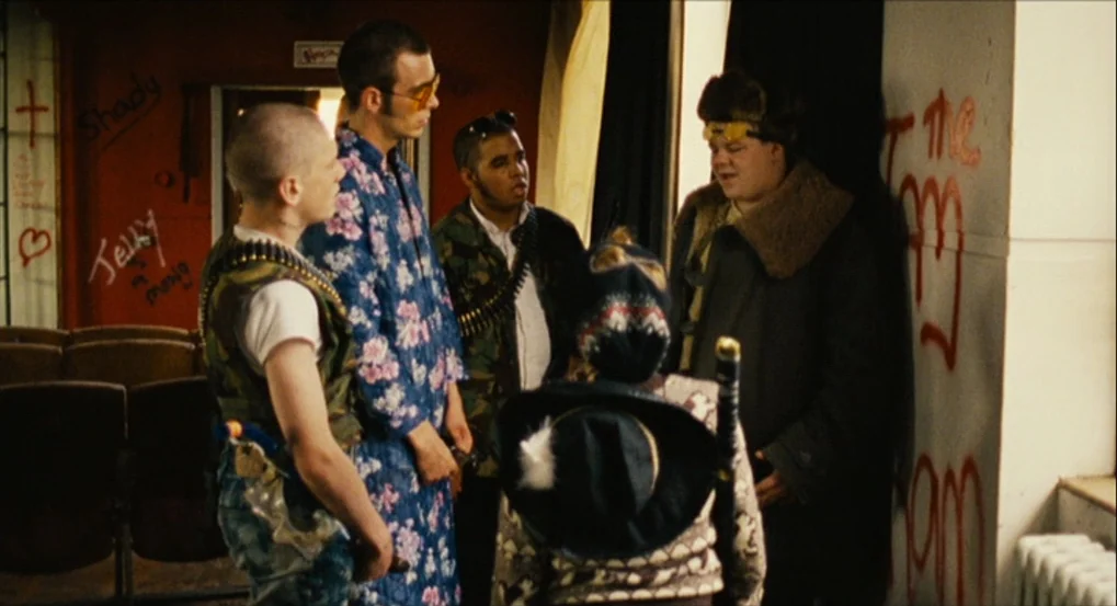 This Is England (2006) movie still: medium — A group of five young men stands in a sparsely furnished room with graffiti. The men wear mismatched…
