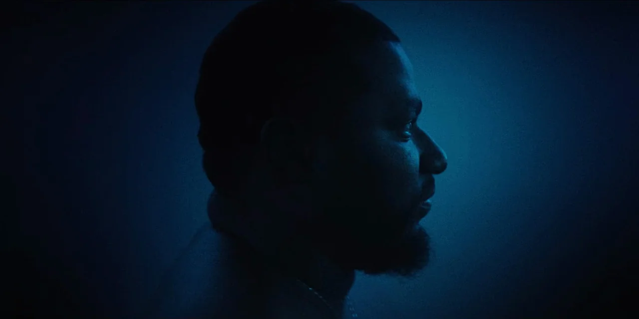 Spermworld (2024) movie still: closeup — A dark, indistinct space with a gradient blue wash across the background. A man in side profile remains…