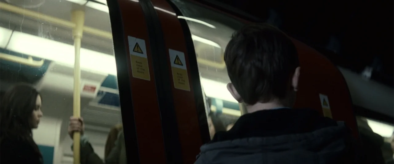 Hereafter (2010) movie still: medium — A dark subway car features red sliding doors marked with hazard warning stickers. A young boy in a hood faces…