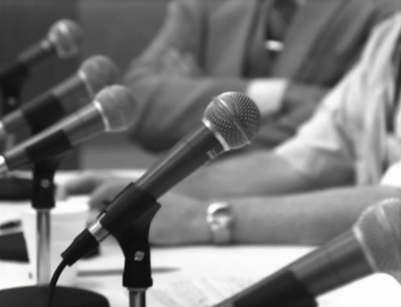 Computer Chess (2013) movie still: medium — A row of multiple microphones sits on a white table at a press event. Partially visible individuals in…