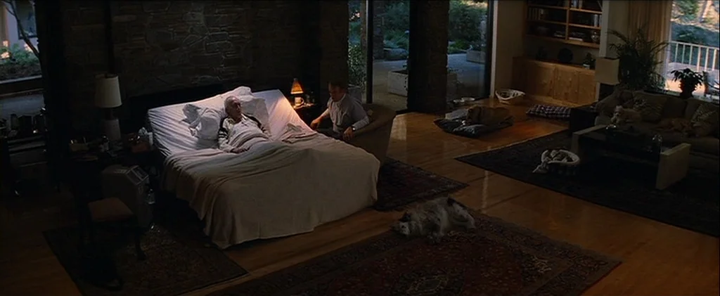 Magnolia (1999) movie still: wide — A spacious modern room features a stone wall behind a medical-style bed. A man sits by the bedside while two…