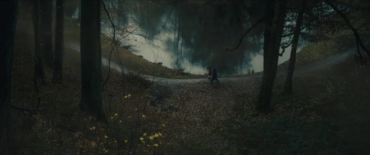 Earwig (2022) movie still: wide — A dark, misty forest landscape features a quiet pond reflecting the tall surrounding trees. Two small figures…