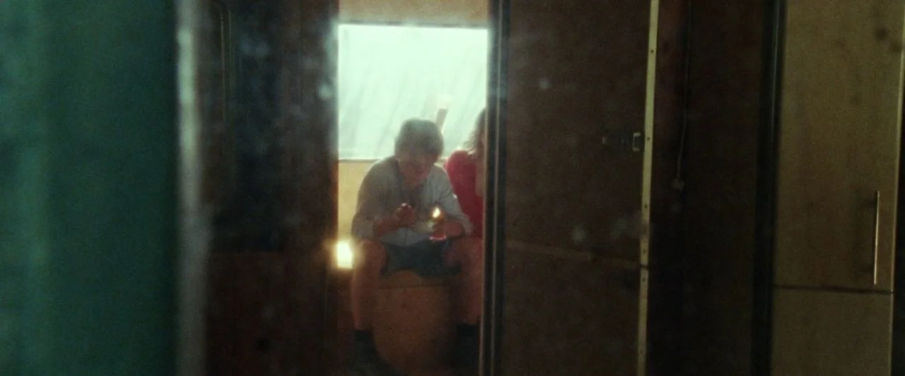 Adoration (2019) movie still: wide — A cramped, dimly lit trailer interior features wooden cabinetry and walls. Two teenagers sit together by a…