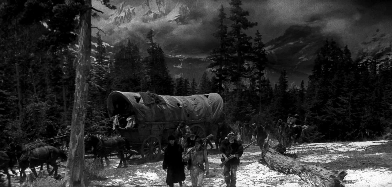 The Big Trail (1930) movie still: wide — A snowy mountain clearing features dense pine forests beneath craggy snow-capped peaks. Men travel on foot…