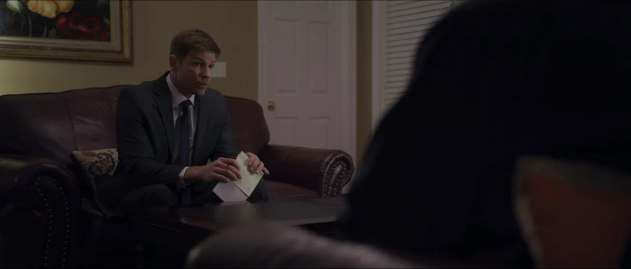 If You're Gone (2019) movie still: medium — A dimly lit living room with dark leather furniture and neutral-colored walls. A man in a suit sits on…