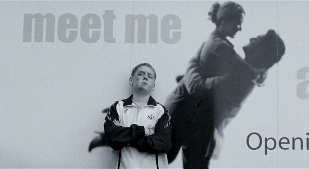 Somers Town (2008) movie still: medium — A man stands before a large billboard featuring text and a couple dancing. The billboard provides a stark…