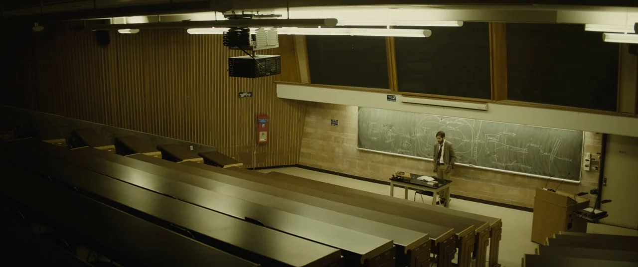 Enemy (2013) movie still: wide — A tiered lecture hall with wooden paneled walls and long horizontal tables. A man stands alone on the stage facing…