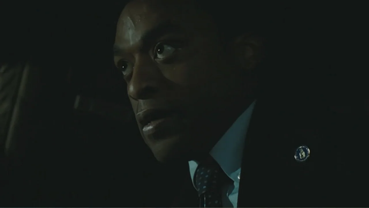 Salt (2010) movie still: closeup — The interior of a dark car at night with minimal ambient lighting. A man in a suit looks toward the passenger…