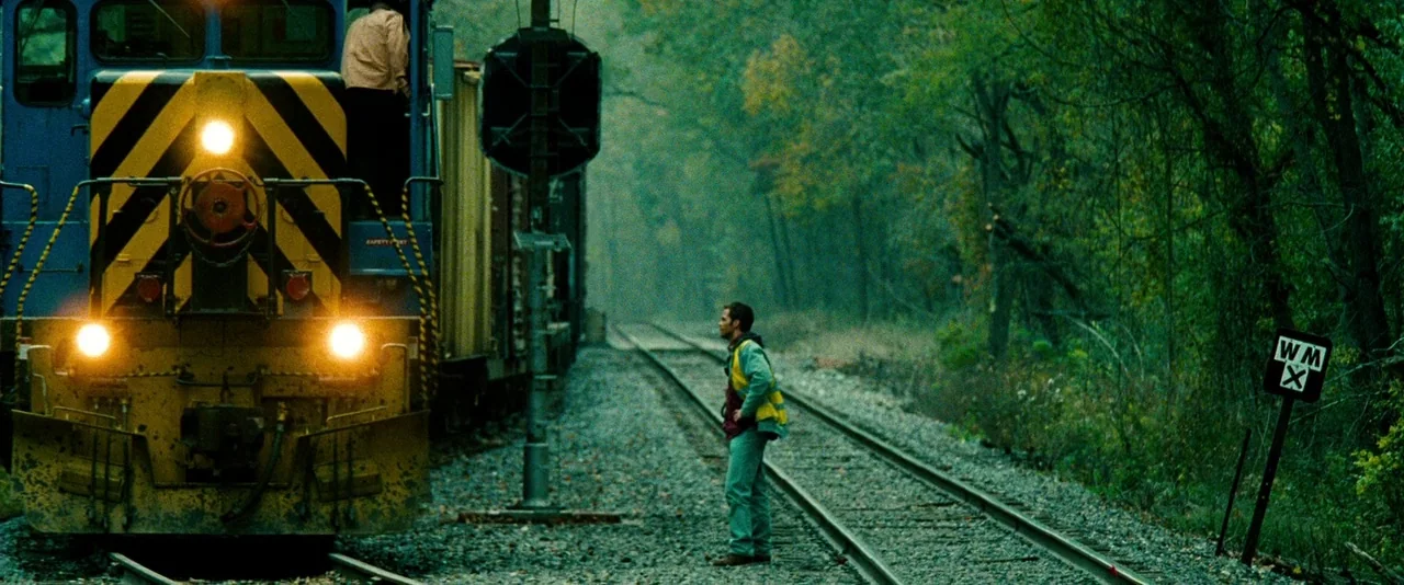 Unstoppable (2010) movie still: wide — A man in a safety vest stands between parallel train tracks in a dense forest. A large locomotive sits idling…
