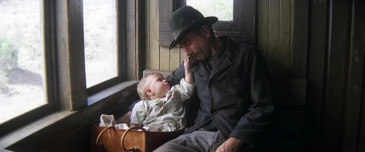 There Will Be Blood (2007) movie still: medium — A man and a small child occupy a wooden seat inside a train carriage. The man leans his head down…