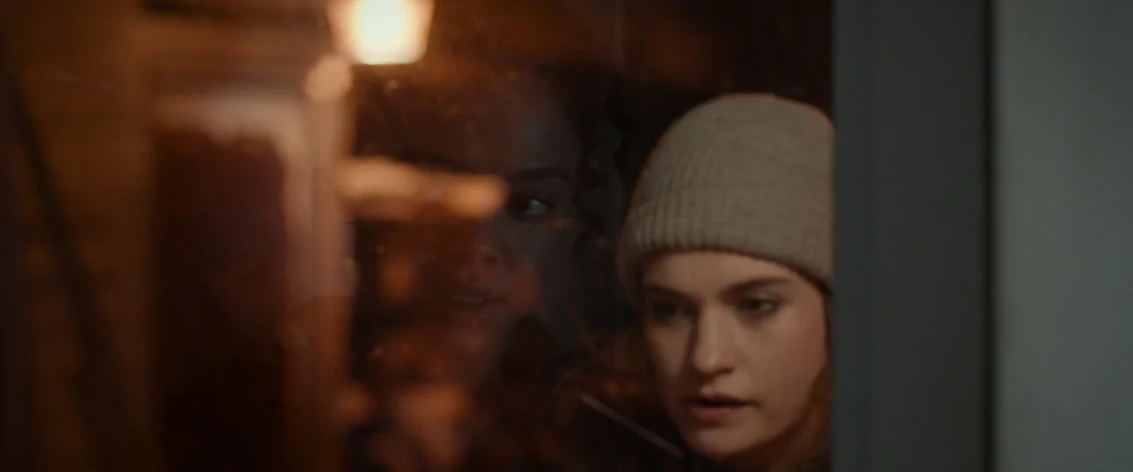 Little Woods (2018) movie still: medium — Two women peer through a vertical glass pane into a dark, out-of-focus area. The woman in the foreground…