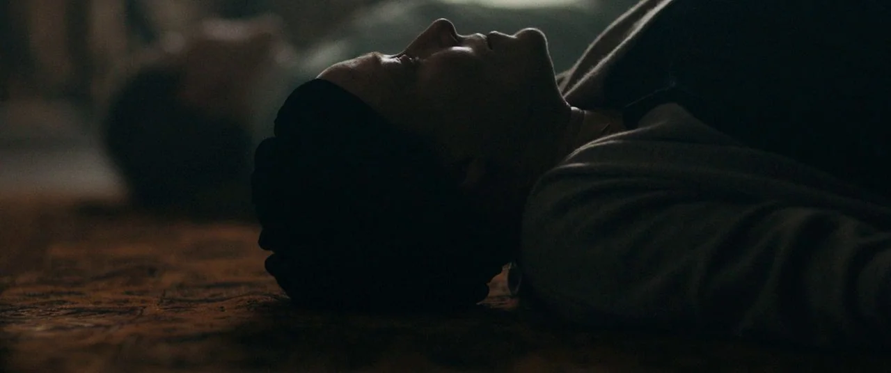 Saint Maud (2019) movie still: closeup — A dark indoor space featuring a textured carpeted floor in low light. A person with closed eyes lies flat…