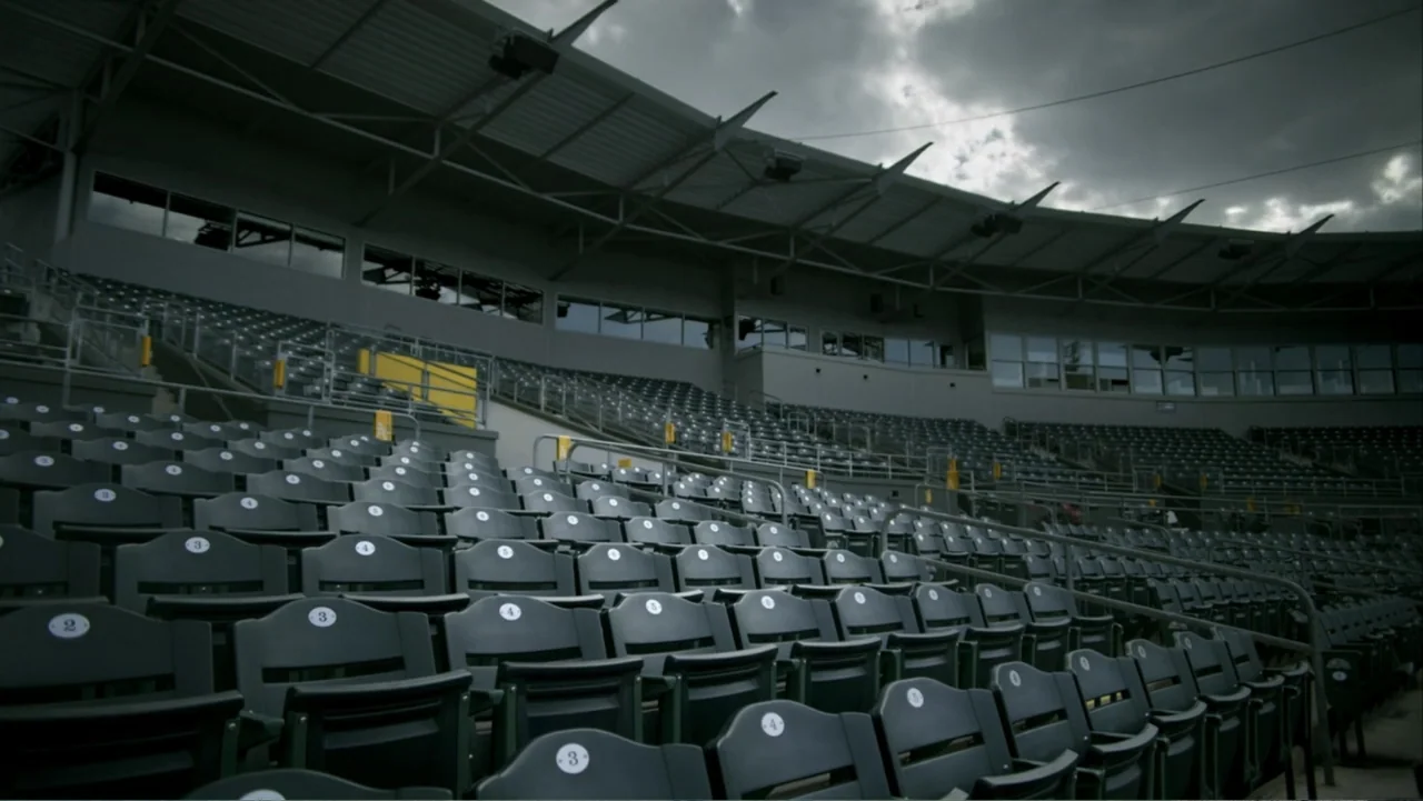 Ferrell Takes the Field (2015) movie still: establishing — An empty outdoor stadium features rows of plastic seats under a heavy overcast sky. The…
