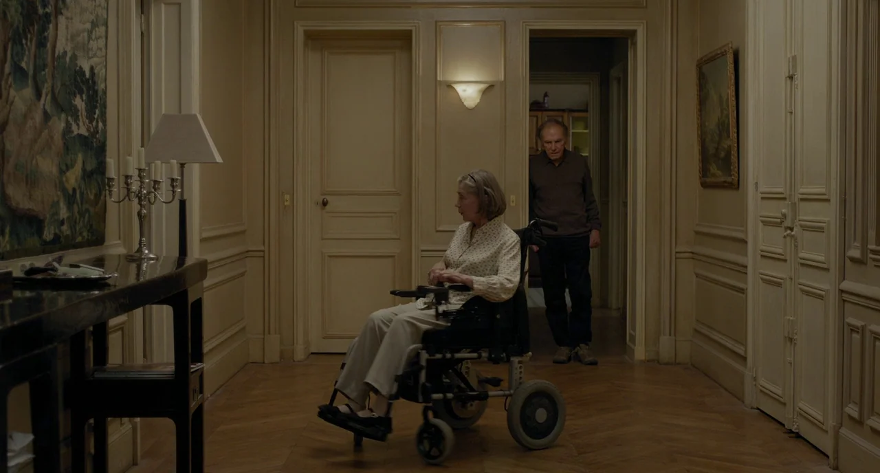 Amour (2012) movie still: fullbody — A refined hallway with beige walls, wooden floors, and framed artwork. A woman in a wheelchair is positioned…
