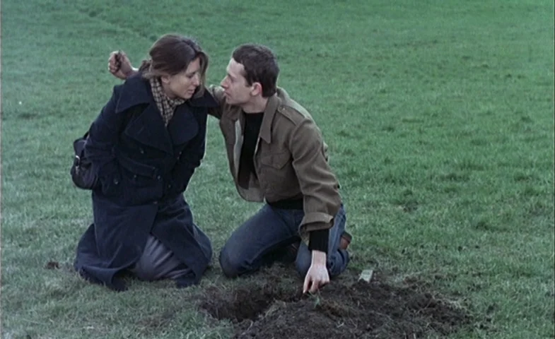 Blind Chance (1987) movie still: fullbody — Two individuals kneel on a grassy field next to an area of disturbed soil. The man touches the dirt…