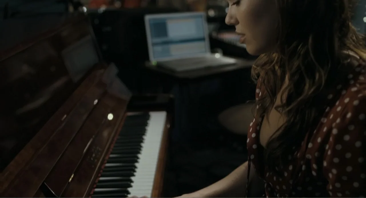 Because I Said So (2007) movie still: medium — A dimly lit recording studio features a wooden piano and an open laptop computer. A young woman with…