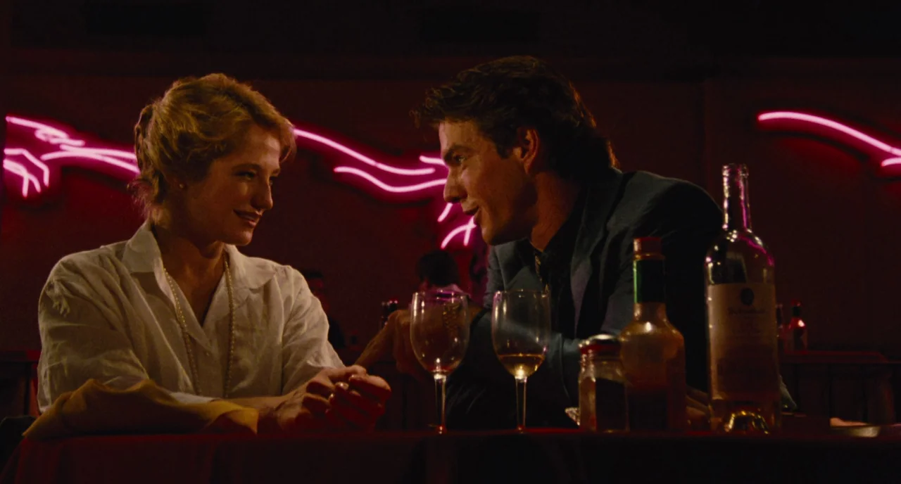 The Big Easy (1986) movie still: medium — A dark bar setting features illuminated neon signage on the back wall. A man and a woman sit at a table…