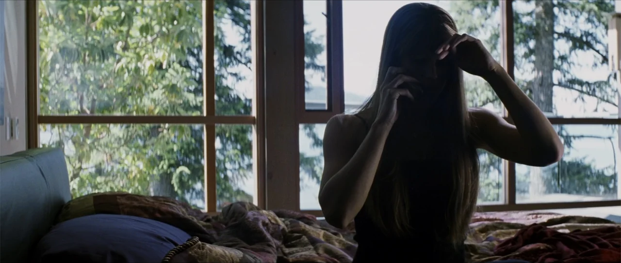 Elektra (2005) movie still: medium — A modern bedroom with a large window wall overlooking dense green trees. A woman sits on a bed with patterned…