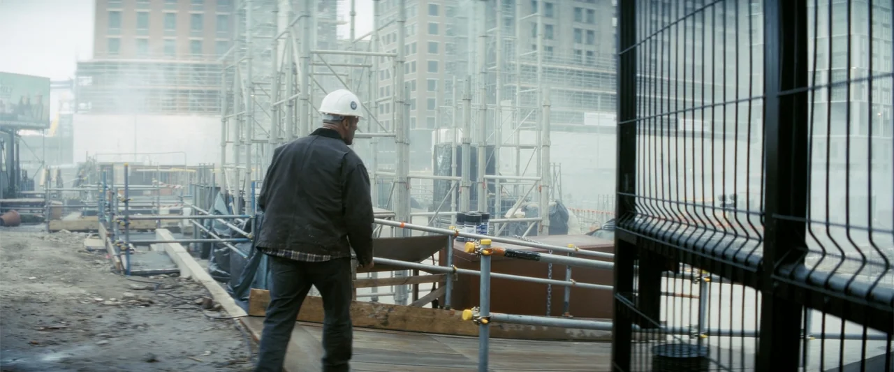 A Working Man (2025) movie still: fullbody — An urban construction site features dense metal scaffolding and hazy, overcast ambient light. A man…